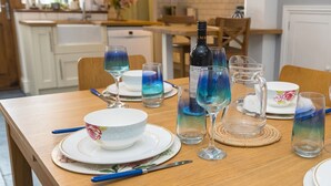 A dining table set with plates, bowls, and glasses, including a bottle of wine, in a well-lit kitchen with wooden cabinets and a window. - Colourful, homely 2 bed terrace - Pass the Keys (Shrewsbury)