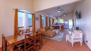 A spacious living area with a wooden dining table, sofa, and a small decorative chair. A kitchen area is visible in the background with a refrigerator and a sink.