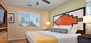 Habitación de hotel con cama, mesita de noche, lámpara, ventilador de techo, ventana con persianas y un cuadro en la pared.