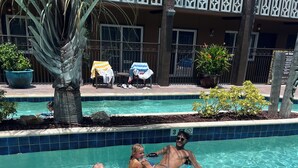 A swimming pool with three people in inflatable tubes, surrounded by a tiled pool edge, a palm tree, and a building with balconies in the background.
