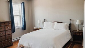 Ein Schlafzimmer mit Holzboden, einem Bett mit weißer Bettwäsche, zwei Nachttischen mit Lampen, einer Kommode und einem Fenster mit blauen Vorhängen.