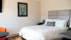 Ein ordentlich bezogenes Bett mit weißen Leinen, ein grau gepolsterter Kopfteil, ein gerahmtes Kunstwerk an der Wand und ein kleiner Tisch mit einer Lampe daneben.