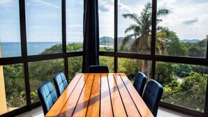 Una mesa de madera con sillas azules en una habitación con ventanas amplias que dan a un paisaje exuberante y una masa de agua.
