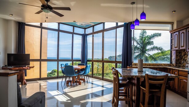Un amplio espacio de estar con un ventanal grande con vistas al océano, un ventilador de techo y una cocina con armarios de madera.
