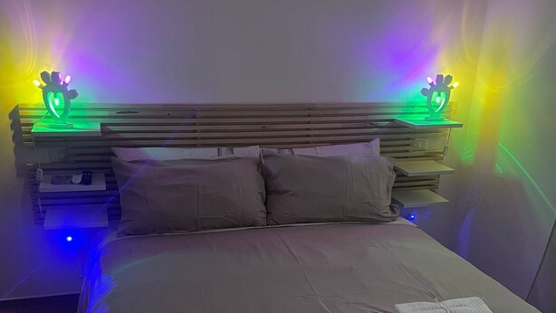 Una cama bien hecha con almohadas y toallas dobladas, una mesita de noche con lámpara, y un banco al pie de la cama.