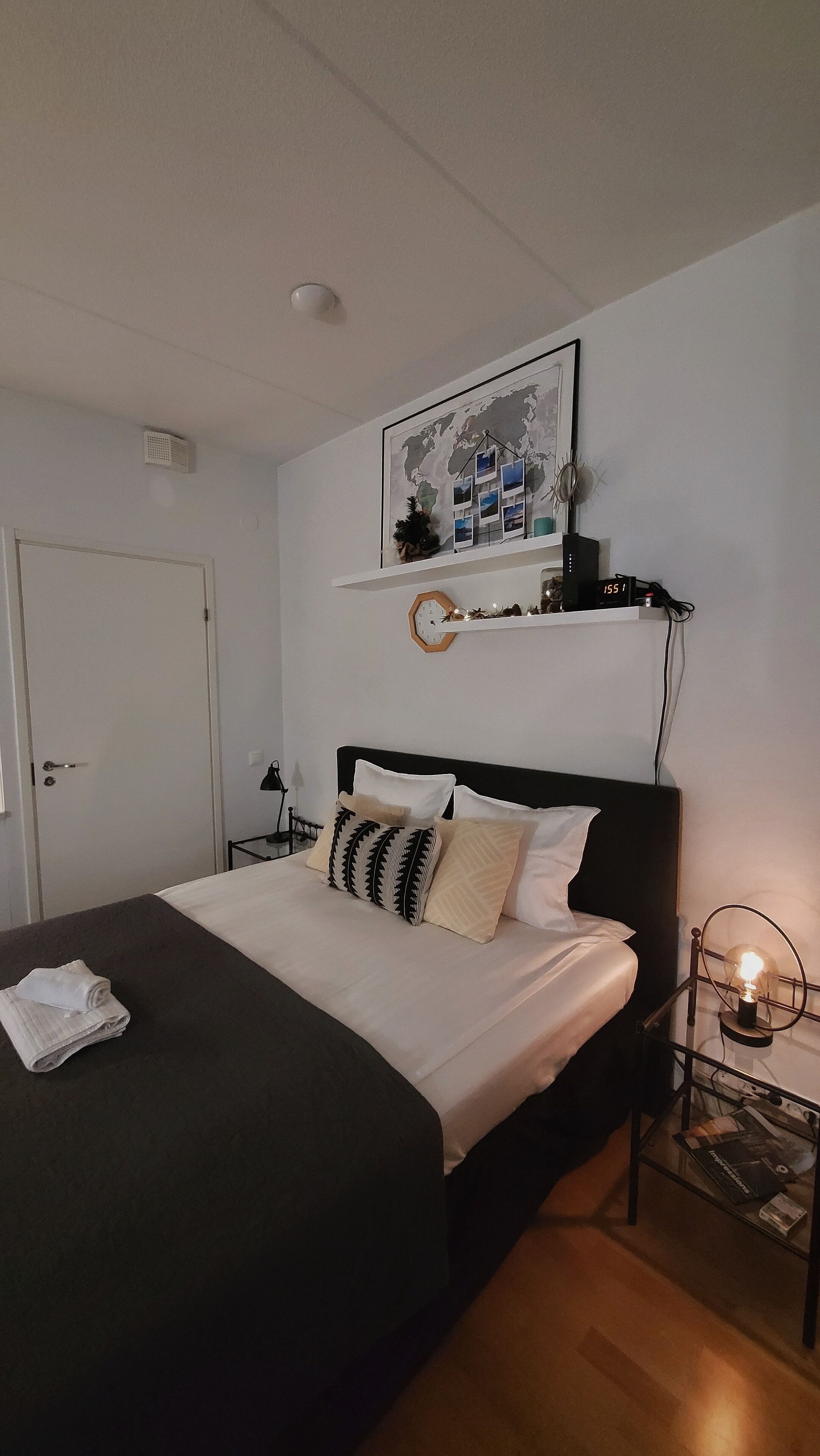 A neatly made bed with a black headboard, a world map artwork above it, and a bedside table with a lamp and a clock.