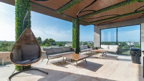 Una terraza en la azotea con sillones colgantes, sofás y una mesa de centro, rodeada de vegetación y amplias ventanas con vistas a la ciudad.
