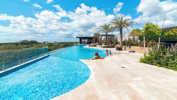 Una gran piscina exterior con borde curvo, rodeada por una zona de patio con sillones de descanso y palmeras.