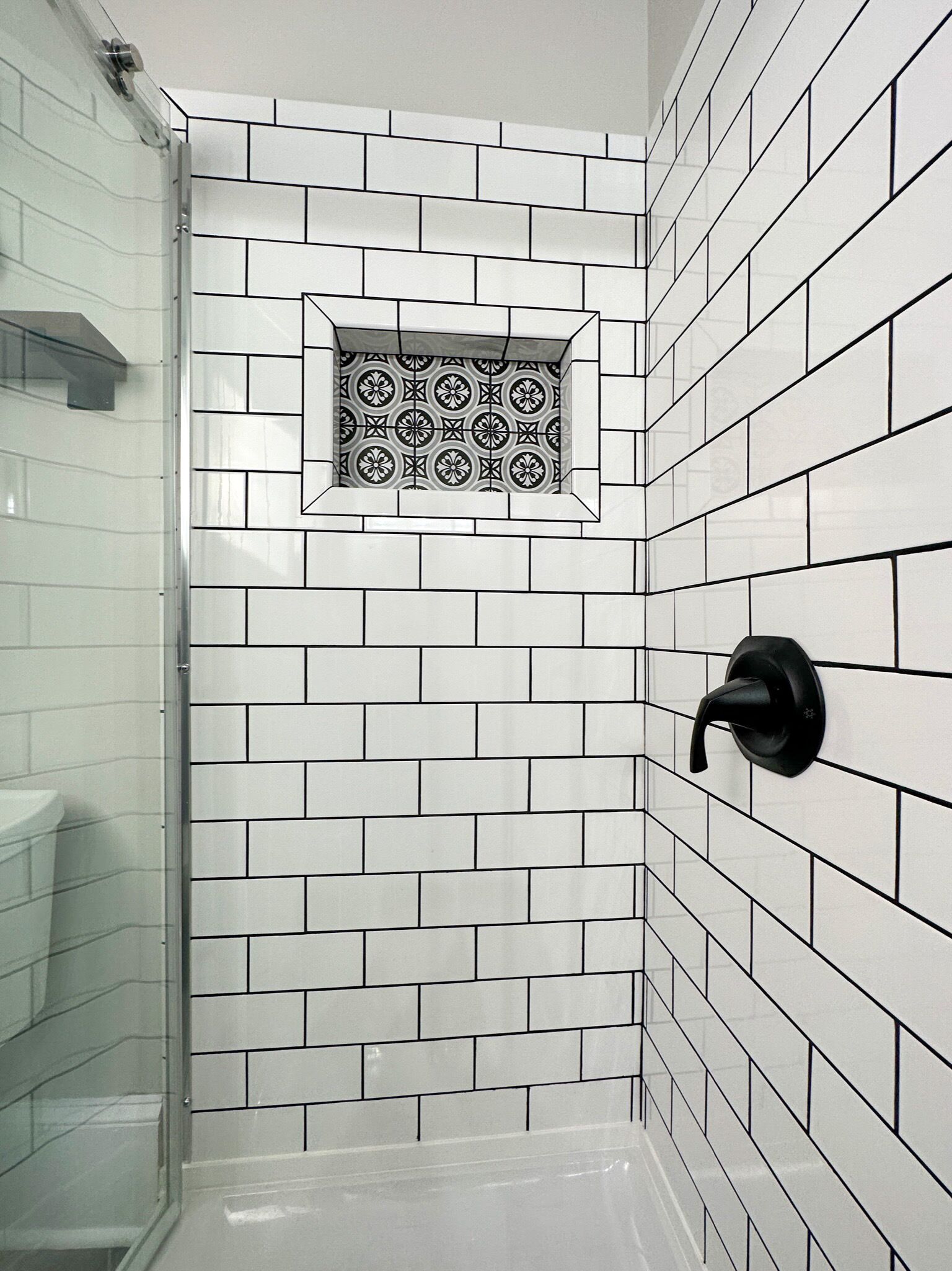 Un baño con azulejos blancos tipo metro, un grifo de ducha negro y un pequeño azulejo decorativo en la esquina.