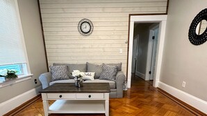 Een woonkamer met een houten visgraatvloer, een witte bank met gekleurde kussens, een salontafel en een wandklok.