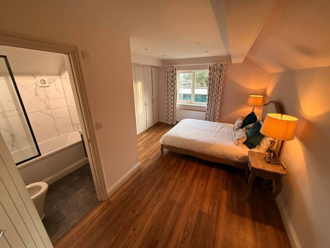 Un dormitorio con cama, mesita de noche con lámpara, ventana con cortinas y vistas al exterior.