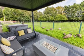 An outdoor patio with a sofa and cushions, a fire feature, and a fenced area with birds. - The Old Coach House: English Cottage with Hot Tub (Hockwold cum Wilton)