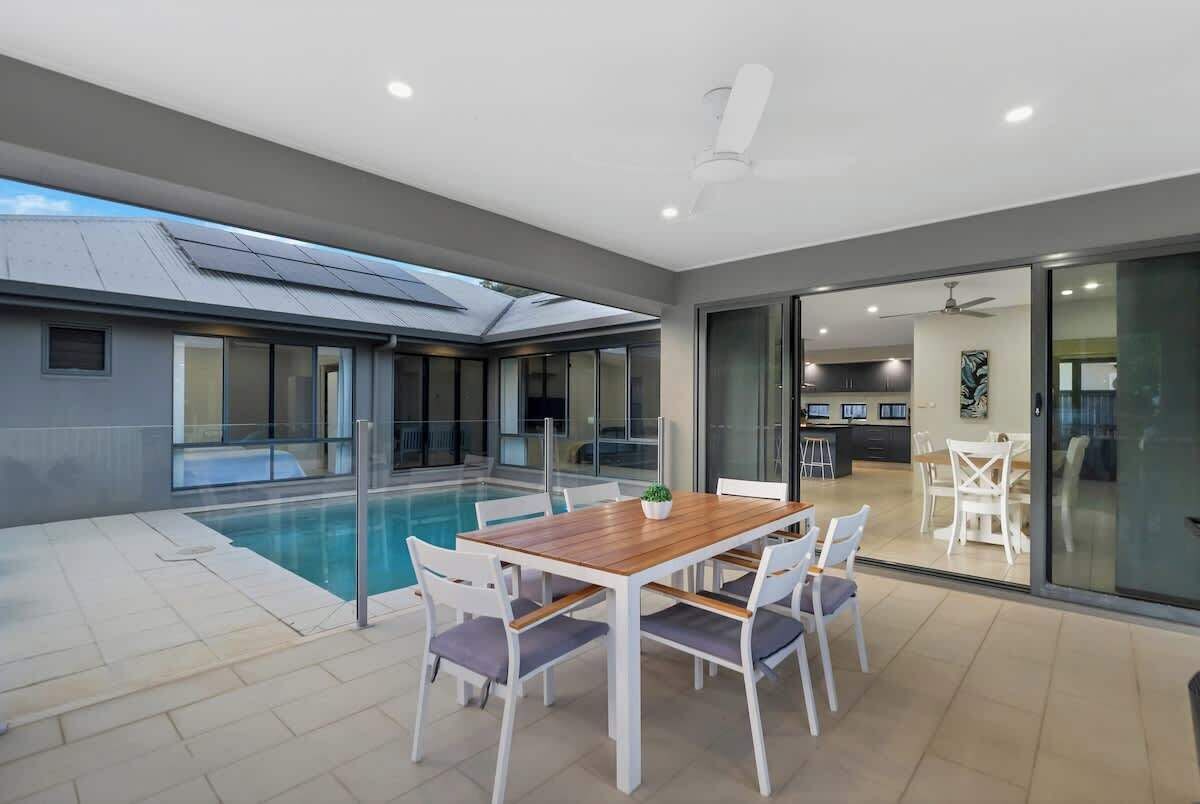 Un espace repas extérieur moderne, doté d’une piscine, d’une table en bois et de chaises blanches.