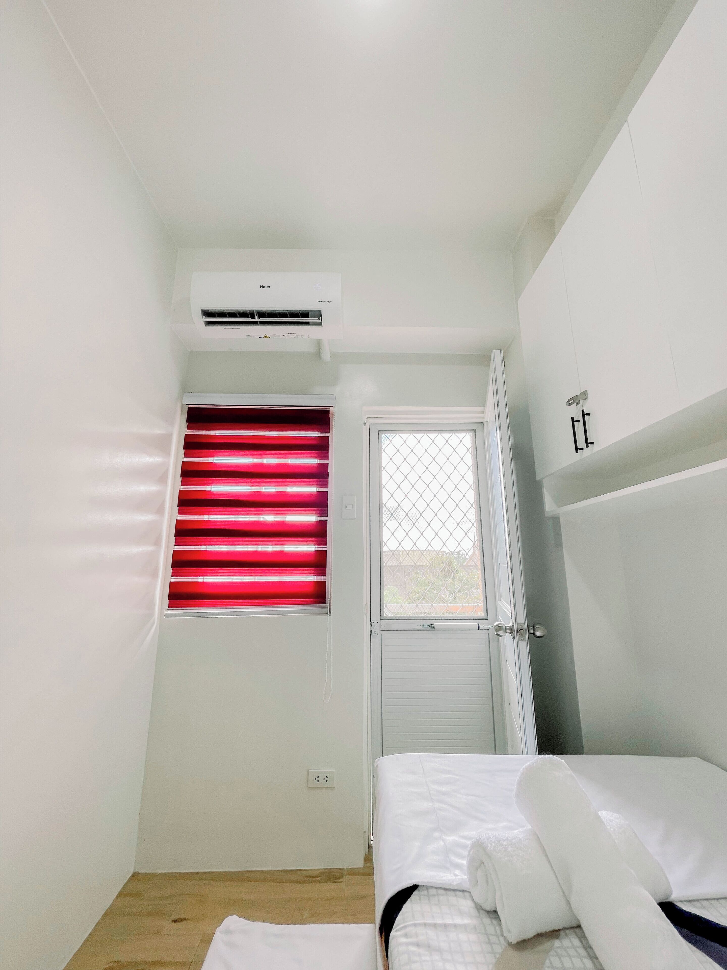 Une petite pièce blanche, avec un lit, une fenêtre avec des stores rouges, un appareil de climatisation et une porte munie d’une moustiquaire.