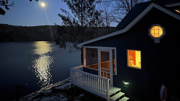 Una cabaña junto a un lago de noche, con luna visible, ventanas y un porche.