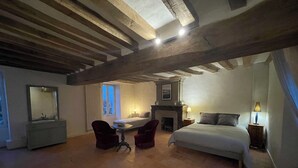 Ein Schlafzimmer mit Bett, Schreibtisch, zwei Stühlen, einem Schminktisch und einem Kamin, sowie einem großen Fenster.