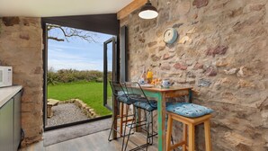 A rustic kitchen with a stone wall, a wooden table, two bar stools, and a view of the garden through an open door.