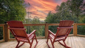 Twee schommelstoelen op een houten terras met uitzicht op bomen en een zonsondergang.