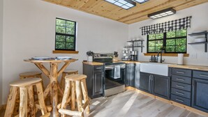 Een keuken met een houten plafond, zwarte kasten, een houten tafel met krukken en grote ramen met uitzicht op groen.