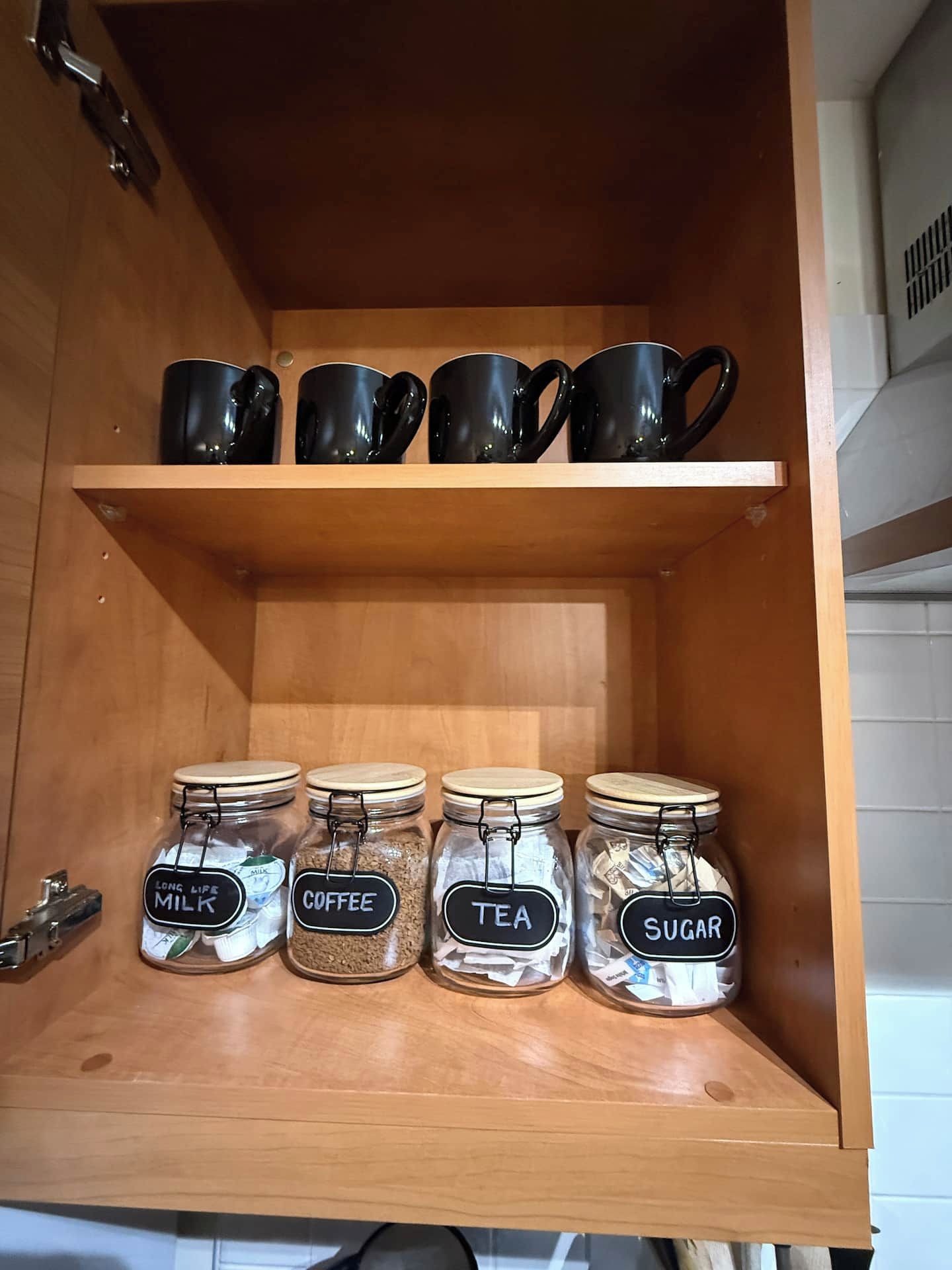 Un armario de cocina con un estante que contiene tazas y tarros negros etiquetados con leche, café, té y azúcar.