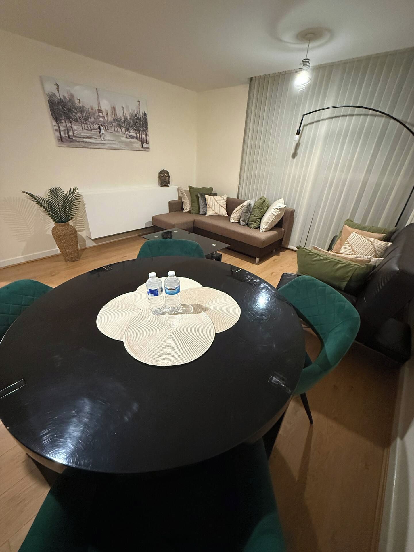 A modern living room with a round black table, green chairs, a sofa with cushions, a potted plant, and a painting on the wall.