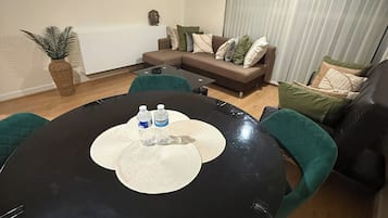 A modern living room with a round black table, green chairs, a sofa with cushions, a potted plant, and a painting on the wall.