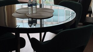 Un moderno salón con una mesa de vidrio, sillas verdes, un sofá con cojines y vistas a un edificio a través de amplios ventanales.