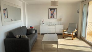 A modern living room with a grey sofa, a white coffee table, a wooden chair, a white cabinet, and a large window with natural light. - CANNES-Vue mer -Superb T2-3 stars -Heating/air-conditioning-Parking (Cannes)
