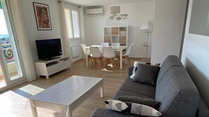A modern living room with a grey sofa, a white coffee table, and a TV on a white stand. There is a dining area with a white table and chairs, and a bookshelf in the background. - CANNES-Vue mer -Superb T2-3 stars -Heating/air-conditioning-Parking (Cannes)