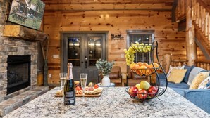 A wooden cabin interior with a stone fireplace, a dining area with a fruit basket, and a bottle of champagne. - Indoor Pool Cabin, Hot Tub & Theater Room, Near Dollywood, Game Room, Sleeps 14 (Pigeon Forge)