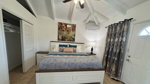 Un dormitorio con cama, mesita de noche, ventilador de techo, un cuadro y una puerta con cortinas.