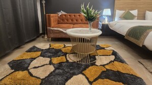 A modern hotel room with a tufted sofa, a round table with a plant, a patterned rug, and a bed with a quilted cover. - Hotel Golden Tulip'z (Gorakhpur)