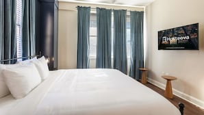 Habitación de hotel con una cama grande, cortinas oscuras, ventilador de techo y un televisor montado en la pared mostrando el nombre del hotel.