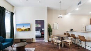 Una sala moderna con un sofá, una mesa de centro y un comedor con sillas. Un cuadro de un castillo adorna la pared, y se ve una cama al fondo.