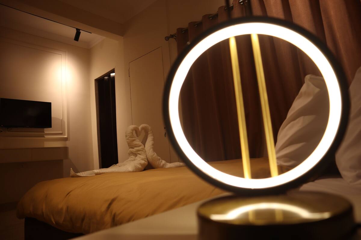 A hotel room with a bed, a television mounted on the wall, and a round mirror with a light on the bedside table.