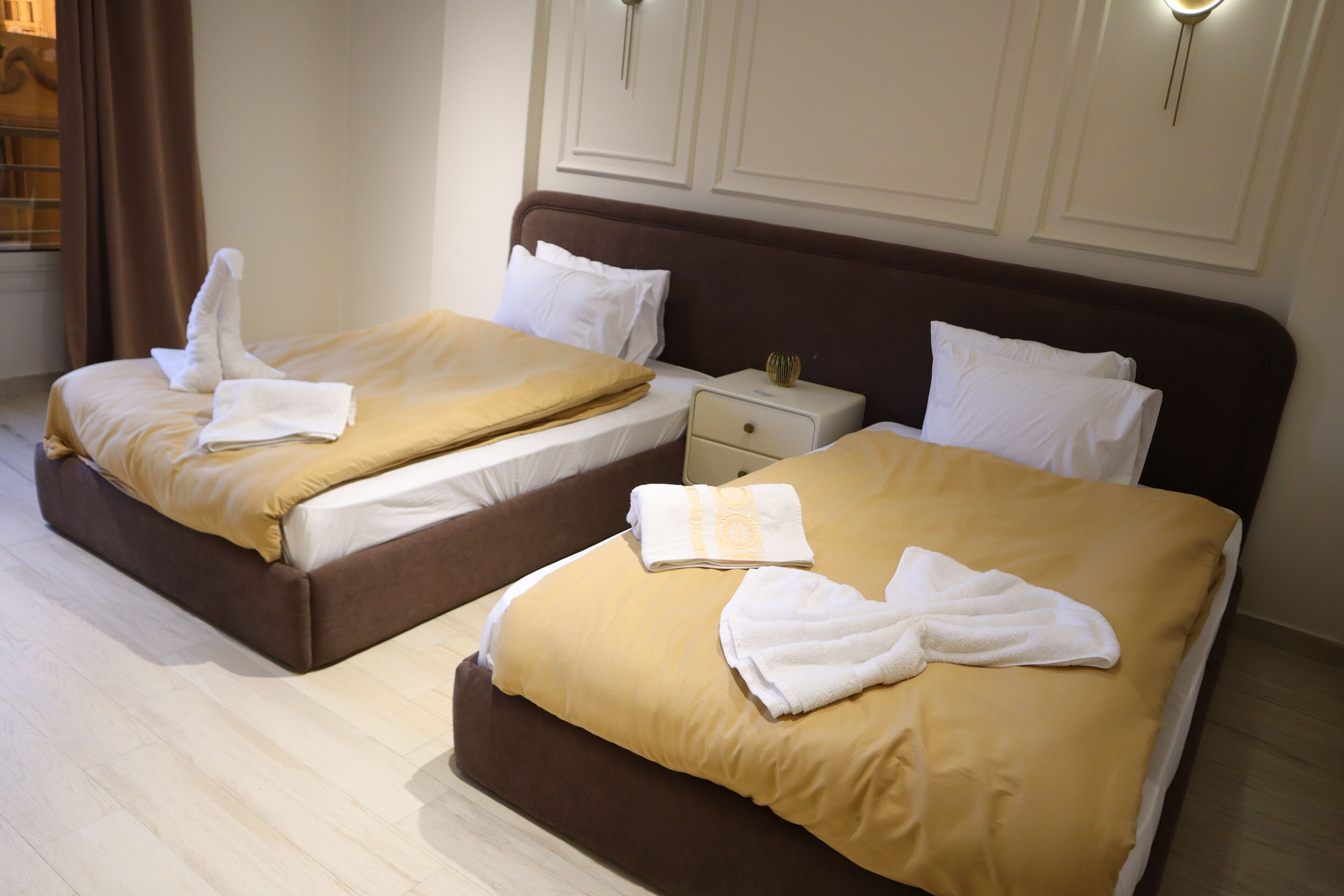 Two double beds with brown headboards and white linens, a nightstand with a lamp, and a window with curtains.