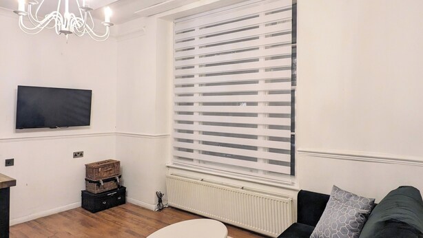 Een woonkamer met een donkere bank, een witte salontafel, een kroonluchter, een televisie aan de muur en een raam met jaloezieën.