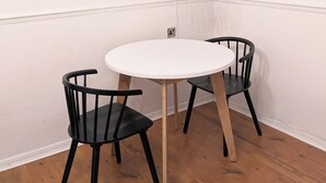 Een kamer met een ronde witte tafel en twee zwarte stoelen. Er is een witte muur met een raam en een hanglamp.
