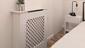 Een slaapkamer met een bed, nachtkastje met lamp, radiator, spiegel en een raam met zonwering.