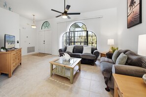 A spacious living room with a ceiling fan, a large window with a view of palm trees, a sofa, a coffee table, and a television on a stand. - 🌴🍹Private Oasis! Heated Pool, Fenced Yard, Poolside Bathroom, Mins to Beach! (Sebastian)