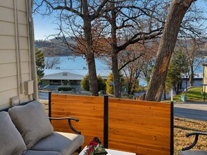 Un banco de madera con cojín, una mesita con un adorno y vista a un lago con edificios al fondo.