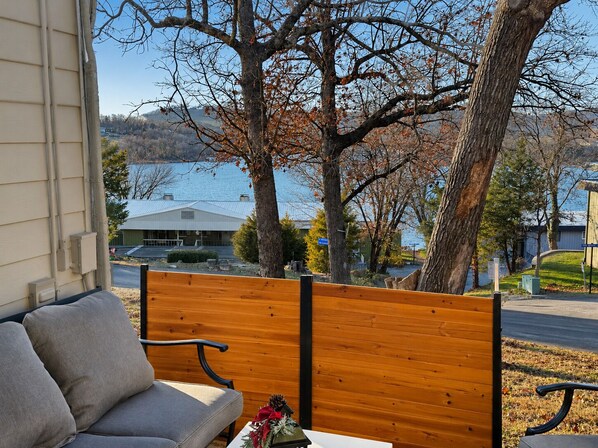 A wooden bench with a cushion, a small table with a decorative item, and a view of a lake with buildings in the background.