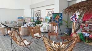 Een kamer met rieten stoelen, een bank en een eettafel. Er staan schilderijen op staffellatten en een rieten dakconstructie.