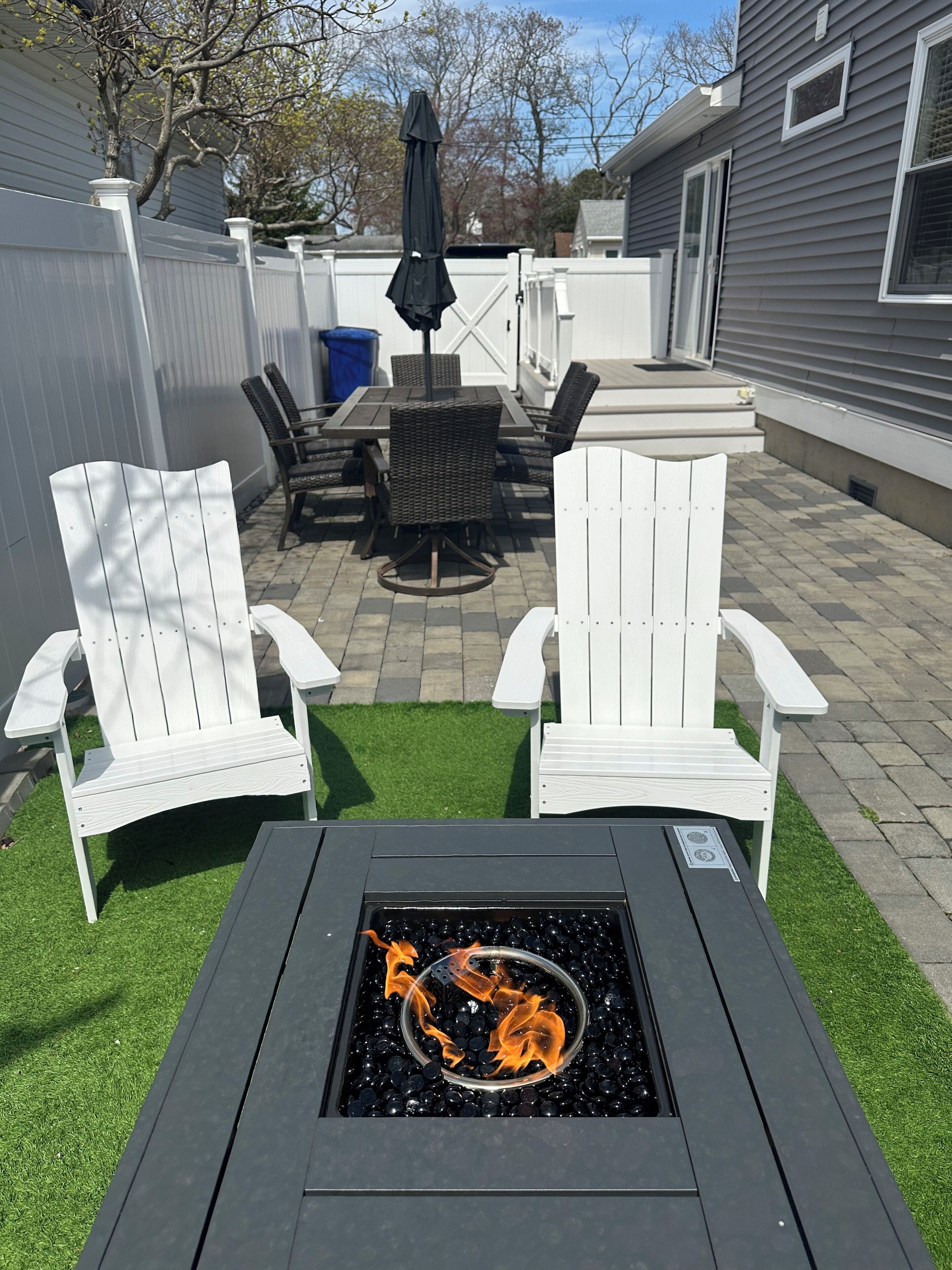 Een achtertuin met een vuurkorf, witte Adirondack-stoelen, een tafel met stoelen en een parasol.