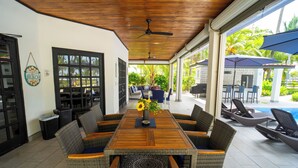 Un espace extérieur abrité, aménagé pour les repas, avec des chaises en osier et une table en bois, offrant une vue sur une piscine et une végétation luxuriante.