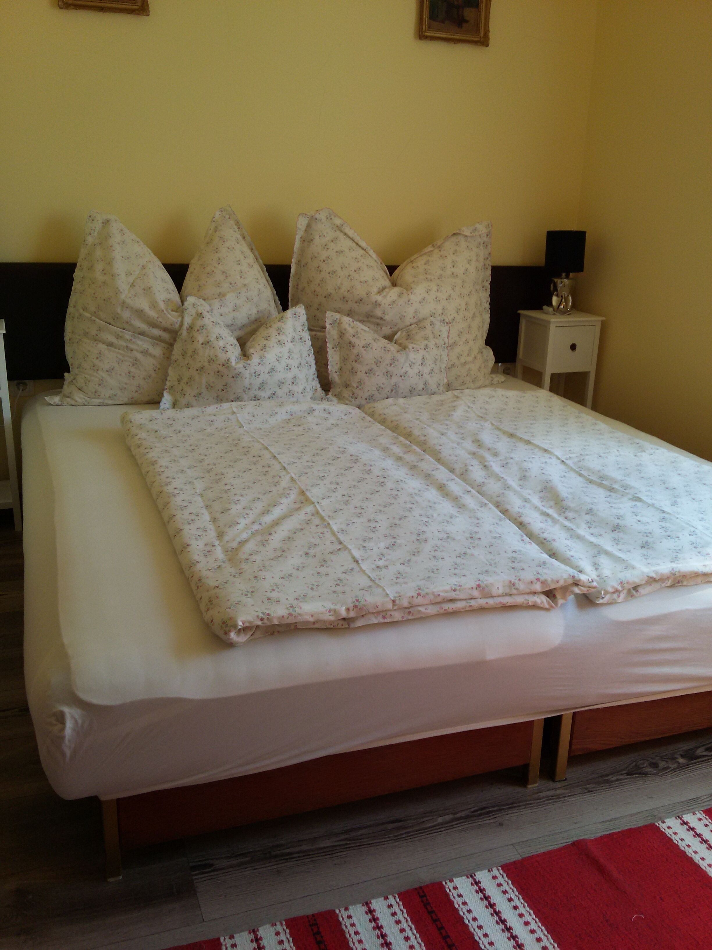 Una cama bien hecha con ropa de cama floral y múltiples almohadas, una mesita de noche con lámpara, y una alfombra a rayas rojas y blancas en el suelo.