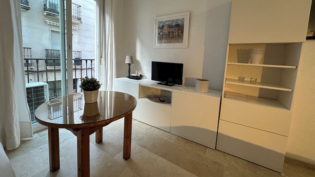 A modern living room with a glass table, a TV on a white cabinet, and a balcony with a view of buildings. - Studio in the center of Malaga (Málaga)