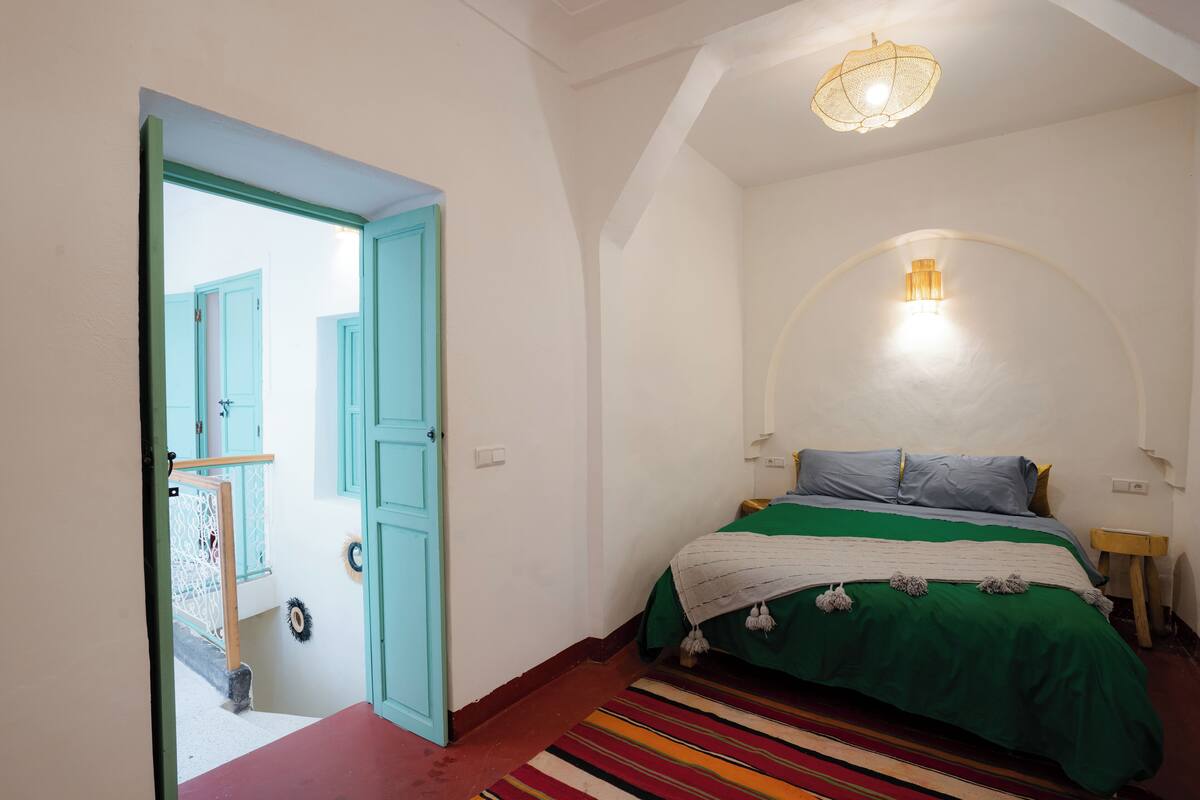 Une chambre à coucher avec un lit, une couvre-lit verte et blanche, un tapis à rayures, un petit tabouret en bois et un luminaire fixé au mur.