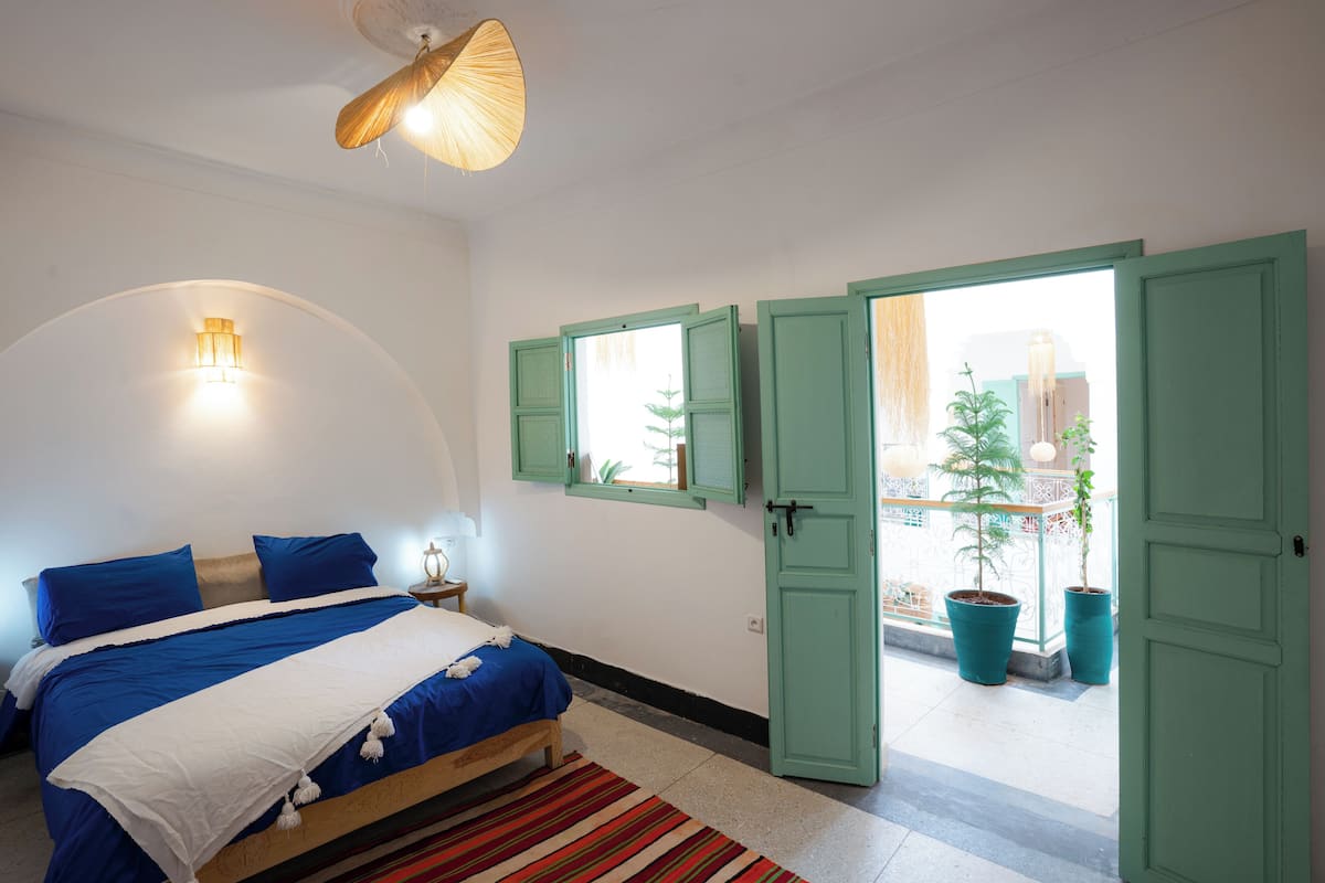 Une chambre à coucher avec un lit, une table de chevet, une lampe, une fenêtre dont les volets sont ouverts, et une vue sur l’extérieur.