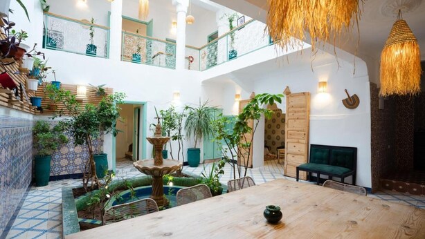 A spacious, multi-level interior with a central wooden dining table, surrounded by chairs. There are multiple balconies with railings, potted plants, and hanging light fixtures. - Riad Lkhlil (Marrakech)